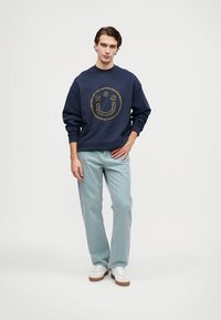 Felpa navy con un sorriso ricamato giallo e testo, abbinata a jeans azzurri chiari, completata con sneakers bianche e suole marroni.