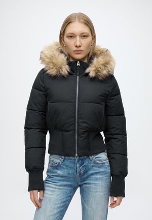 Kvinde iført sort dunjakke med pelskantet hætte og blå jeans, stående foran en ensfarvet lys baggrund og kiggende fremad.