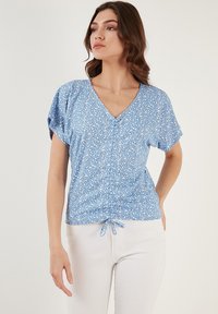 Blusa floreale blu con maniche corte, scollatura a V, dettagli arricciati in vita e un tessuto morbido e leggero. Indossata con pantaloni bianchi.