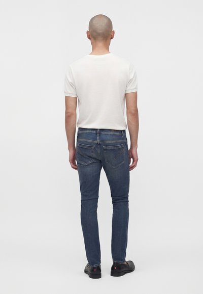 Homme portant une chemise blanche à manches courtes unie, un jean bleu ajusté et des chaussures noires, debout, tourné de dos, sur un fond blanc.