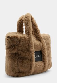 Faux fur totebag in lichtbruin met een pluizige textuur. Voorzien van een zwart logo-embleem, dikke handvatten en een gestructureerde vorm.
