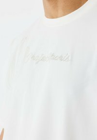 Camiseta de algodón blanca con cuello redondo y bordado "projectparis" en un sutil beige, con una textura suave y lisa.