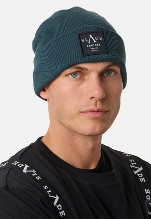 Knit beanie in dark teal with ribbed texture and a square black patch displaying "SLADE COUTURE." Model wears a black t-shirt with white accents.
