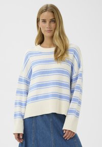 Jeune femme aux longs cheveux blonds portant un pull blanc à rayures horizontales bleu clair et une jupe en denim bleu, debout devant un fond blanc.