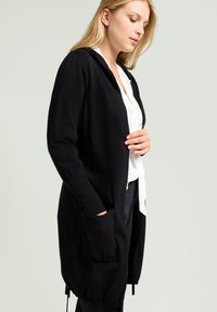 Long gilet noir avec capuche, fabriqué dans un tissu doux. Il possède de grandes poches avant et un ourlet à cordon de serrage, porté sur un chemisier blanc.