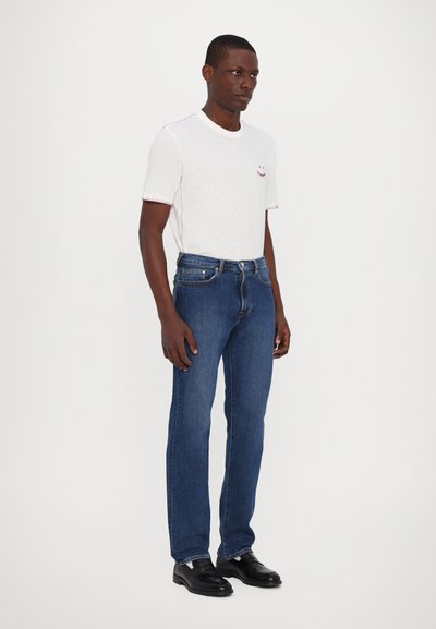 T-shirt blanche à manches courtes avec des détails de surpiqûres rouges, accompagnée de jeans bleus à jambe droite et de chaussures en cuir noires. Design simple et minimaliste.