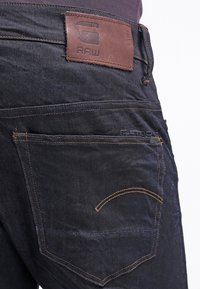 Vaqueros de mezclilla azul oscuro con un parche de cuero marrón etiquetado como "RAW" en la parte posterior. Presentan costuras amarillas y un diseño distintivo en el bolsillo trasero.