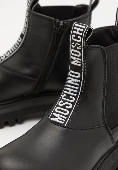 Botas de tobillo de cuero negro con paneles laterales elásticos y una tira de tirón blanca con la marca. Presenta una suela gruesa y textura suave. Cierre con cremallera.