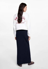 Bílý zkrácený svetr s červeným potiskem na zadní straně, v kombinaci s dlouhou námořnickou sukní z texturovaného materiálu.