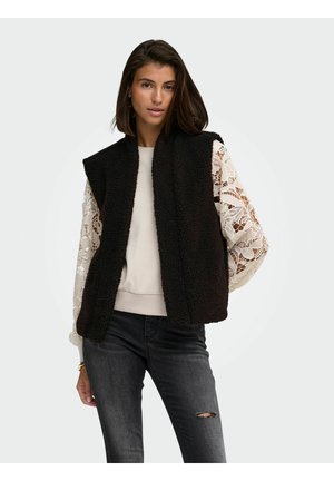 Kvinde iført sort ærmeløs fleecevest over hvid blonde langærmet top og mørke revnede jeans mod ensgrå baggrund.