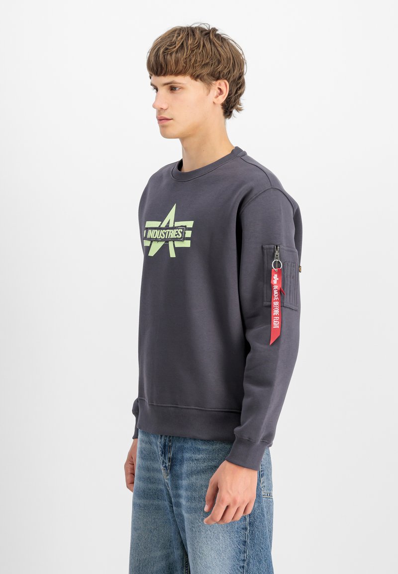 Sötétszürke pulóver, kerek nyakkal, lime zöld logóval és cipzáras zsebbel a bal ujján, piros textilcímkével kiemelve.