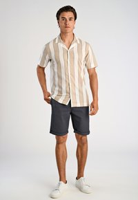 Man met een beige en witte verticaal gestreepte korte mouwen shirt, donkere opgerolde shorts en witte sneakers, staand tegen een effen achtergrond.