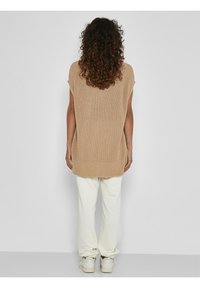 Beige pletený oversized top s krátkými rukávy, kombinovaný s krémovými tepláky a bílými teniskami, pohled zezadu. Hladká textura.