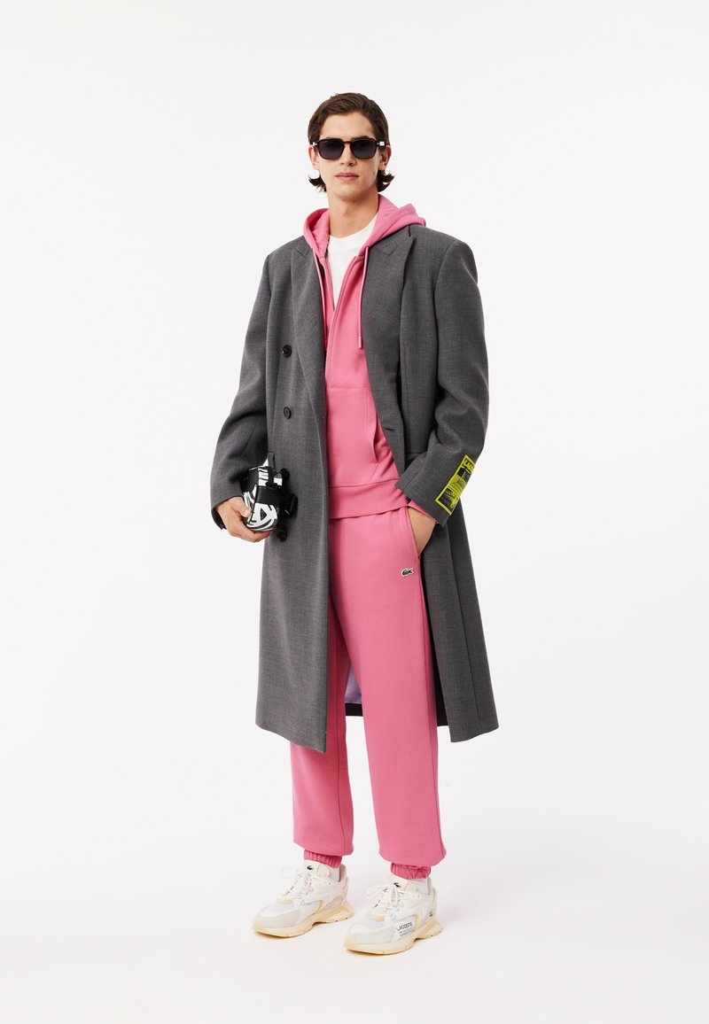 Cappotto grigio doppiopetto su una felpa rosa con cappuccio e pantaloni coordinati. Il modello indossa sneakers bianche e grigie, tenendo una borsa nera.