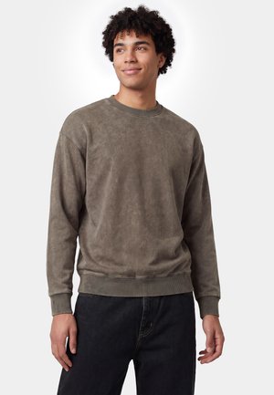 Junger Mann mit lockigem Haar, der einen bräunlich-grauen langärmeligen Pullover und schwarze Hose trägt, steht vor einem schlichten, hellen Hintergrund.