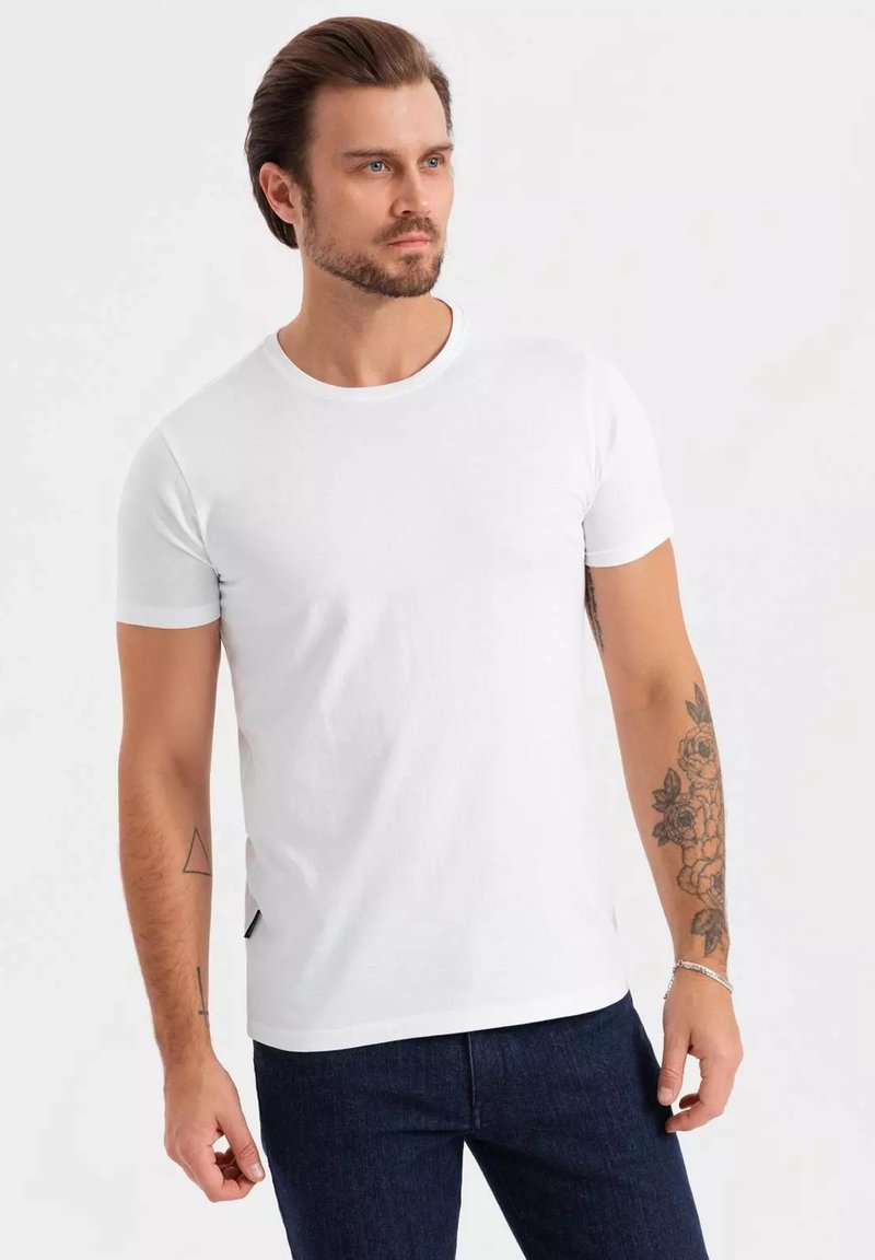 Weißes kurzärmliges T-Shirt aus Baumwolle mit Rundhalsausschnitt. Getragen mit dunkelblauen Jeans; tattoo am linken Arm sichtbar.