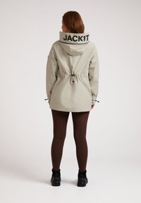 Lichtgrijze jas met verstelbare trekkoord in de taille, grote capuchon en "JACK1T" logo op de rug. Draag het met een paar bruine leggings en zwarte schoenen.