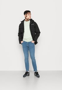 Jovem com vitiligo vestido com uma jaqueta preta, um suéter verde claro, jeans azuis e ténis pretos, de pé com as mãos nos bolsos, olhando para o lado.