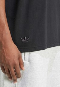 Black cotton t-shirt featuring a small black embroidered trefoil logo on the hem, paired with light gray shorts.