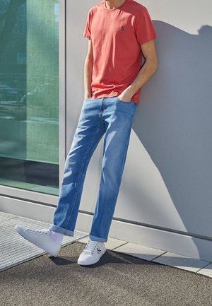 Camiseta roja de manga corta, jeans azul claro y zapatillas blancas. Tela texturizada, jeans de corte recto y puños remangados en los zapatos.