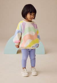 Spalvingas, per didelis megztinis su pasteliniais banguotais raštais; derinamas su šviesiai mėlynais ribotais tampriais ir baltais sportbačiais; fone neutrali spalva.