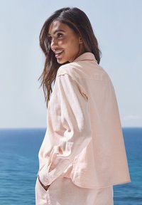 Light pink, long-sleeve shirt with a relaxed fit, tailored collar, buttoned cuffs, and a soft, textured fabric, set against a blue ocean background.