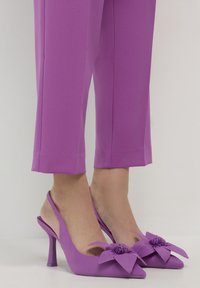 Scarpe slingback viola con tacco alto, punta affusolata e grandi decorazioni floreali, indossate con pantaloni viola a 3/4.