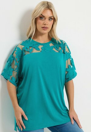 Kvinde med blondt hår iført en turkis top med gennemsigtigt blomsterblonde på skuldre og ærmer, poserer mod en hvid væg.