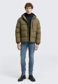 Olijfgroene pufferjack met ritssluiting, geribde mouwen en capuchon. Gecombineerd met blauwe jeans en zwarte enkellaarsjes. Glad, gewatteerd textuur.
