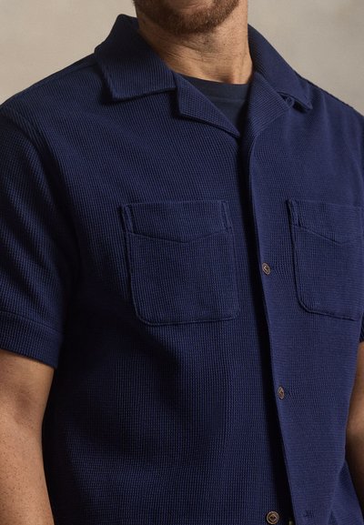 Μπλε ναυτικό κοντομάνικο πουκάμισο από υφασμένο ύφασμα με ραβδωτό μοτίβο, με γιακά, δύο τσέπες στο στήθος και κλείσιμο με κουμπιά.