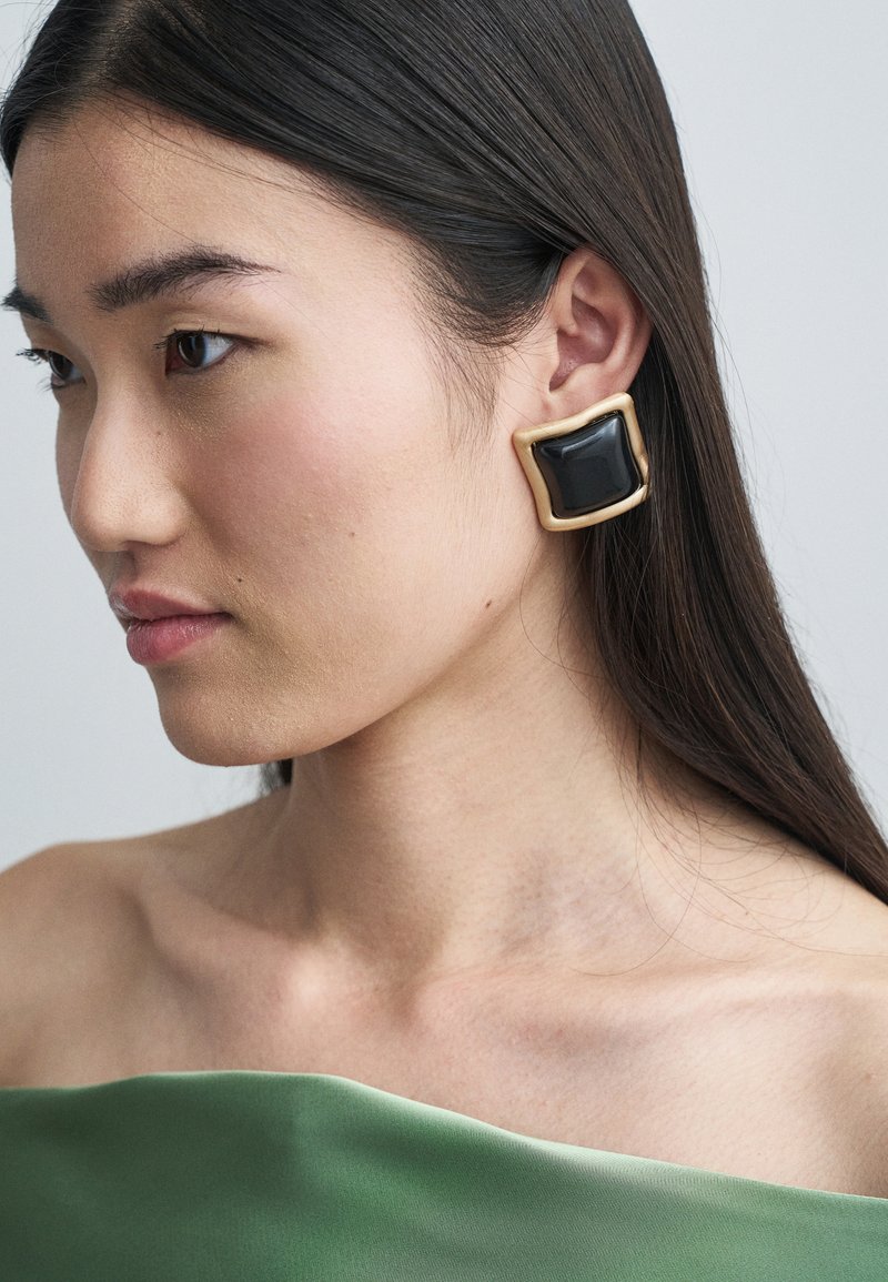 Black and gold square-shaped earring featuring a glossy texture, worn on the left ear against a light background, complementing a green top.