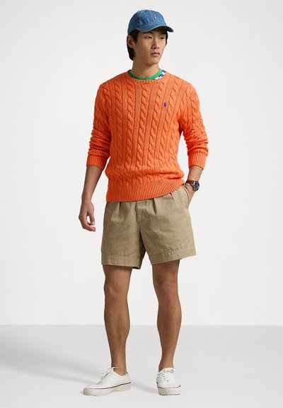 Orange cable-knit sweater, khaki shorts, and white sneakers. Completing look with a denim cap. Simple, casual outdoor outfit.