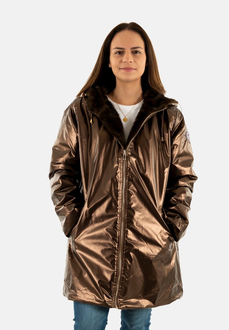 Giacca lunga in metallo bronzeo con colletto in pelliccia sintetica, zip frontale e tasche laterali, abbinata a jeans blu e una camicia bianca sotto.