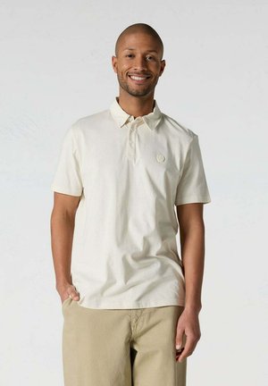 Hombre sonriente con cabello corto lleva un polo de manga corta color marfil y pantalones beige, de pie relajado frente a un fondo liso y claro.