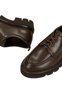 Een paar donkerbruine leren veterschoenen met dikke zwarte robuuste rubberen zolen op een witte achtergrond.