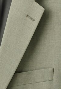 Primo piano del risvolto di una giacca di abito verde kaki che mostra una asola e una tasca a filetto con tessuto finemente strutturato.