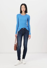 Lange mouw geribbelde top in helderblauw, met een gegolfde ronde halslijn, gecombineerd met donkere skinny jeans en witte sneakers. Bruine schoudertas inbegrepen.