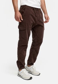 Pantalones cargo de pana marrón con puños elásticos, bolsillos laterales y cintura con cordón, combinados con zapatillas grises y blancas.