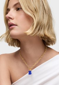 Collar de cadena de oro con un charm de oso texturizado azul, presentado sobre un fondo claro con una blusa de hombros descubiertos.