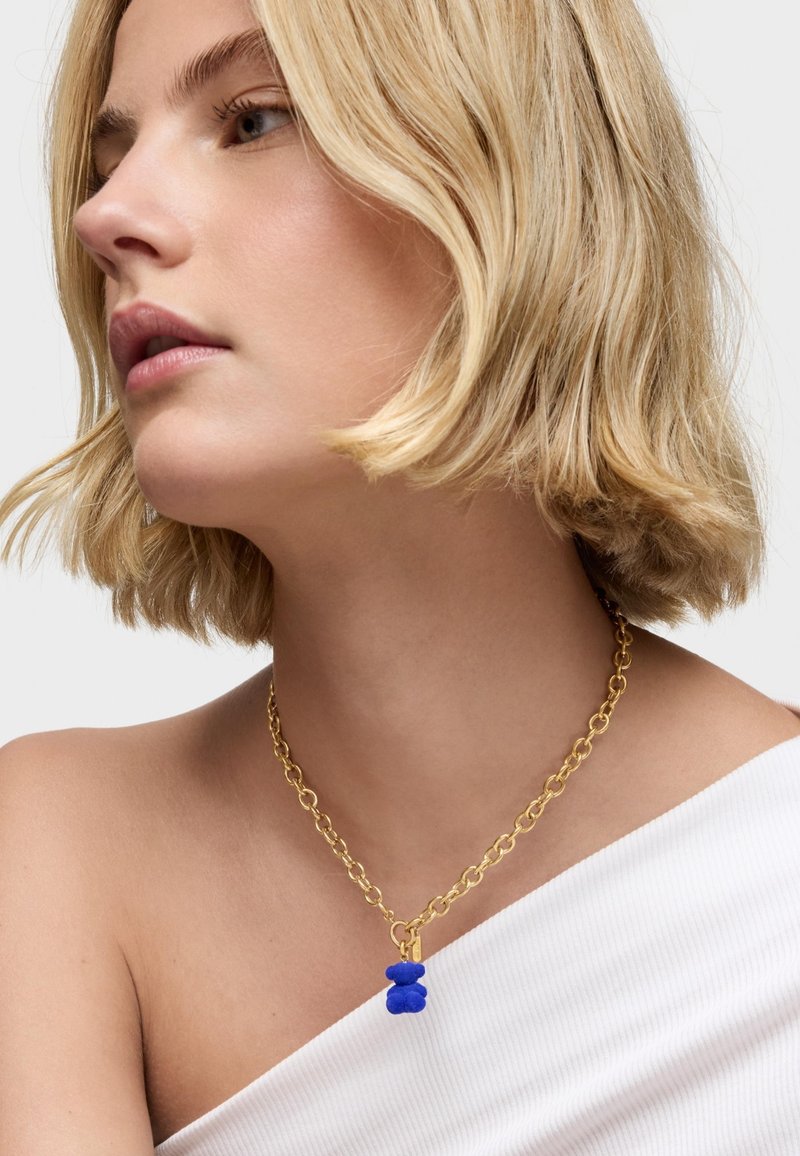 Collar de cadena de oro con un charm de oso texturizado azul, presentado sobre un fondo claro con una blusa de hombros descubiertos.