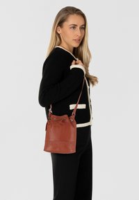 Braune Leder-Eimer-Tasche mit Zugbandverschluss, geprägtem Logo und silberner Hardware, die an der Schulter einer neben einem Modell im schwarzen Pullover getragen wird.