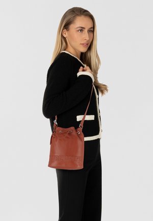 Borsa a secchiello in pelle marrone con chiusura a coulisse, logo impresso e hardware argentato, indossata a spalla accanto a un modello in un maglione nero.