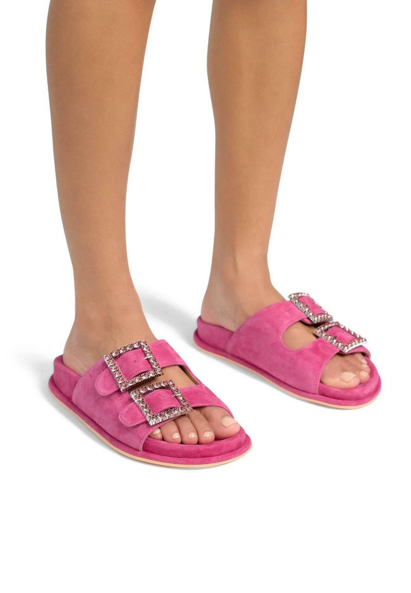 Sandales à glissière en daim rose, ornées de deux grandes boucles décoratives avec des strass. La semelle est plate avec un design arrondi.