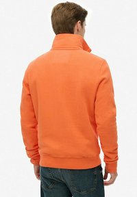 Orange sweatshirt with a high collar and ribbed cuffs, featuring a smooth texture and relaxed fit. Back view shows minimal detailing.