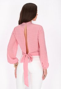 Rosa strukturierte Bluse mit langen Ärmeln, offenem Rücken und Bindeband in der Taille. Das Material scheint leicht und weich zu sein.