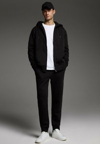 Czarna bluza z zamkiem, spod której wystaje biała koszulka, w połączeniu z czarnymi dresami i białymi sneakersami. Gładki materiał, casualowy styl.