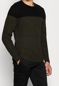 Stickad tröja med svart överdel och olivgrön kropp, med ett texturerat mönster och ribbad halsringning. Långa ärmar, avslappnad passform.