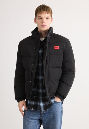 Young man wearing a black puffer jacket with a red "HUGO" patch, a black and blue plaid shirt, and blue jeans, standing against a plain background.