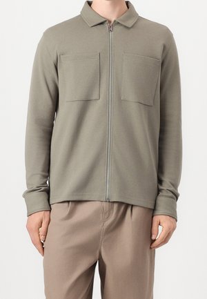 Man wearing olive green long-sleeve zip-up shirt with two chest pockets, paired with light brown pants, standing against a white background.