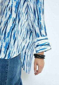 Camicia texturizzata blu e bianca con un motivo a strisce verticali, caratterizzata da un polsino arrotolato e un orlo arrotondato, abbinata a denim.